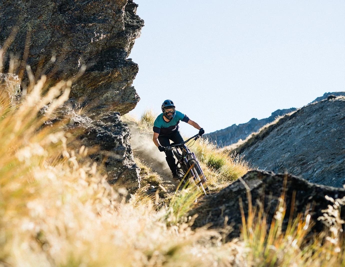 Mountain biker riding down a narrow, rocky trail at the Cardrona Bike Park surrounded by tall grass and steep cliffs on a sunny day.