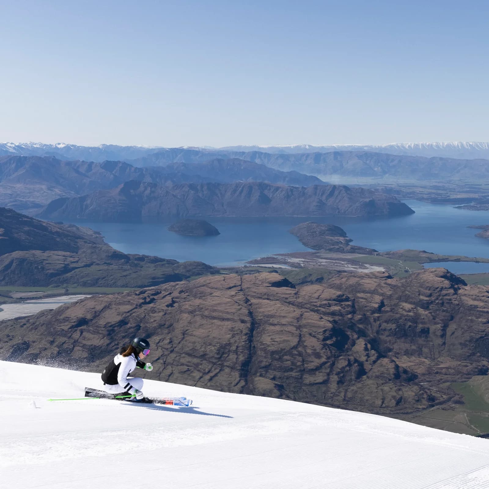 A skier races down the pristine slopes of Treble Cone, with a stunning view of Lake Wānaka and the surrounding mountains in the background, under a clear blue sky.