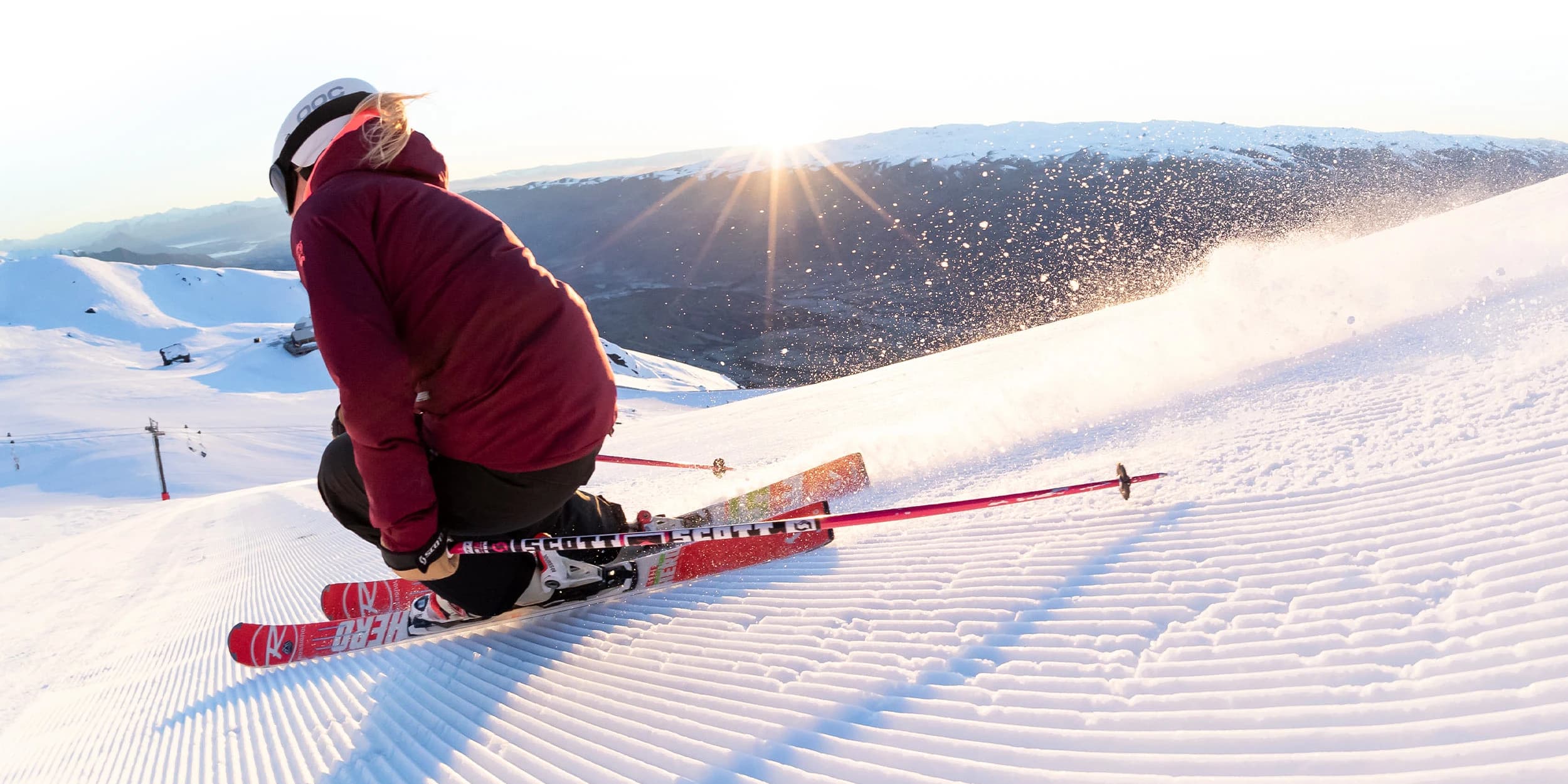 A skier in a maroon jacket carves on a snow-covered mountain at the top of a ski slope during sunset. The skier is wearing a white helmet and goggles and has red skis. The scene is illuminated by the soft golden sunlight casting a warm glow over the freshly groomed slope, with distant mountains fading into the background under a clear sky.