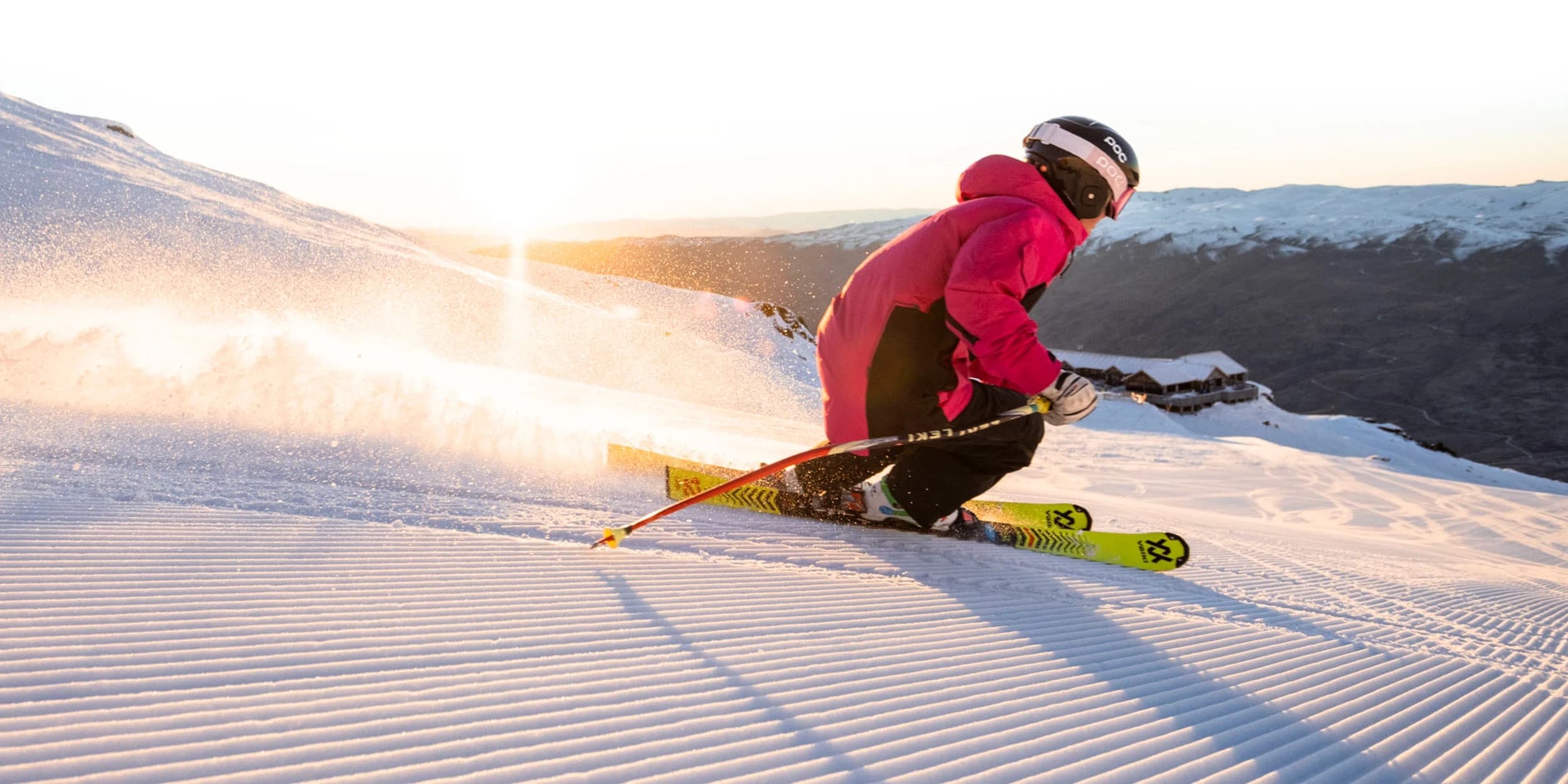 A skier in a bright pink jacket glides down a groomed slope at sunrise, kicking up snow with the mountains in the background and golden light illuminating the scene.