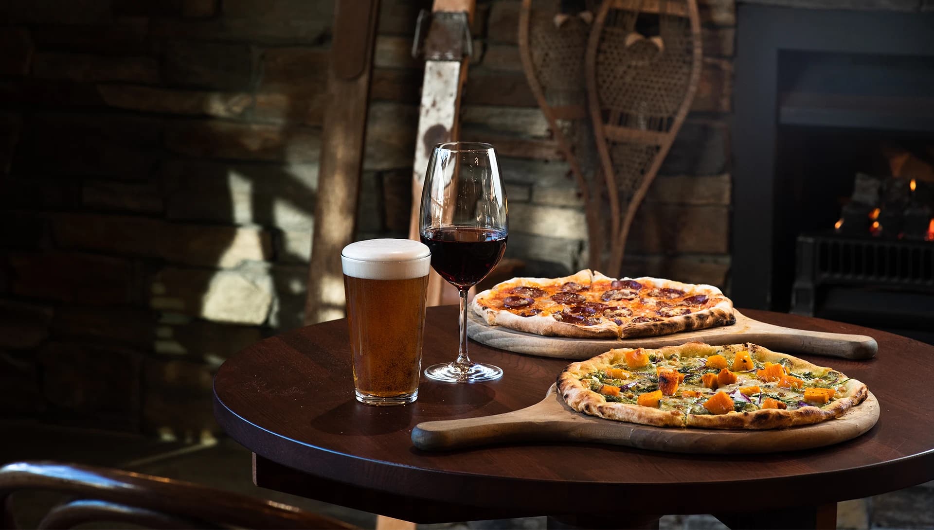 Two wood-fired pizzas on boards, a glass of red wine and a pint of beer sit on a table in a lounge area with a lit fireplace in the background.