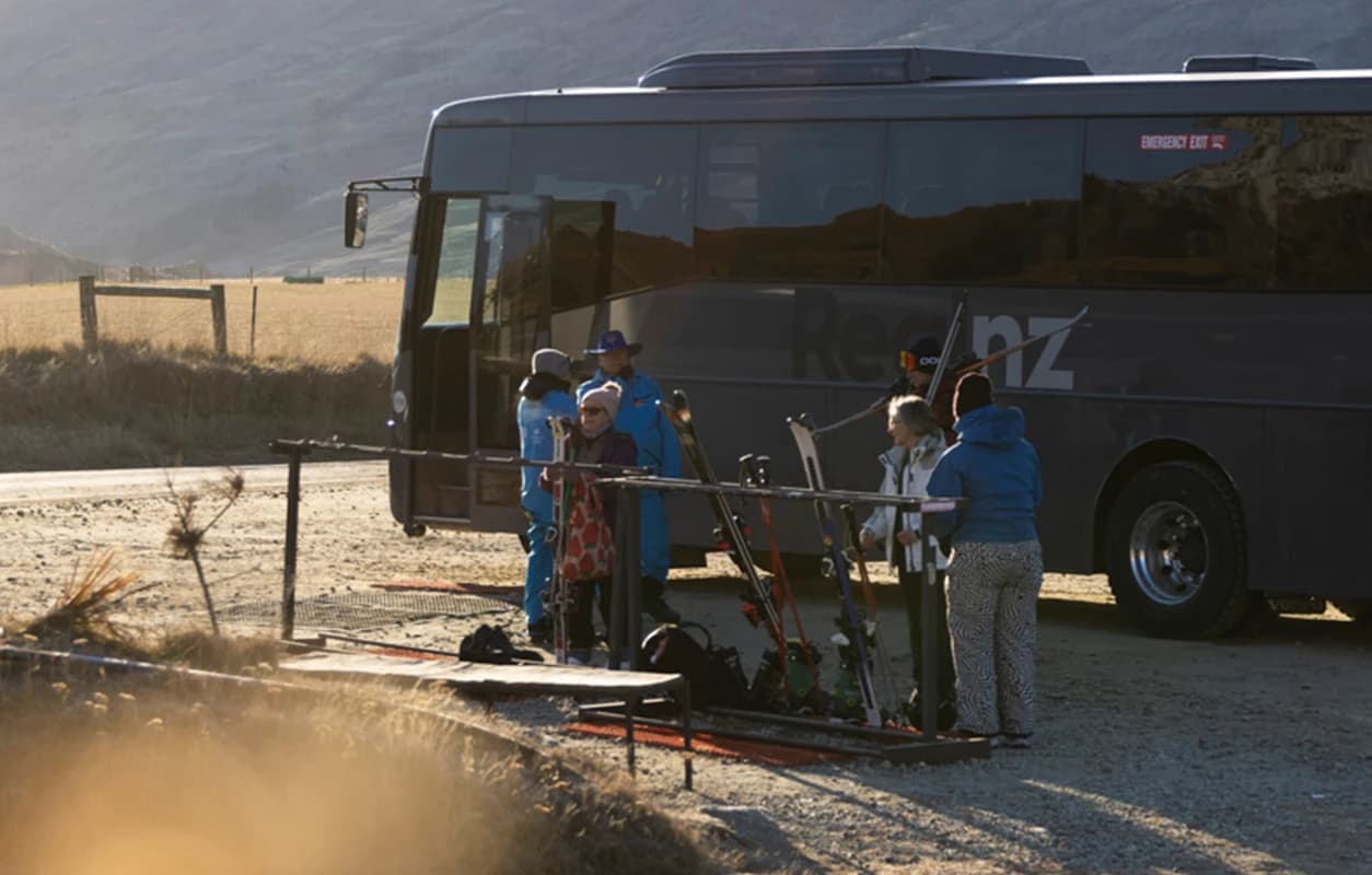 A group of skiers gathers beside a bus, preparing their gear near a ski rack, with golden fields and distant mountains under soft sunlight in the background.