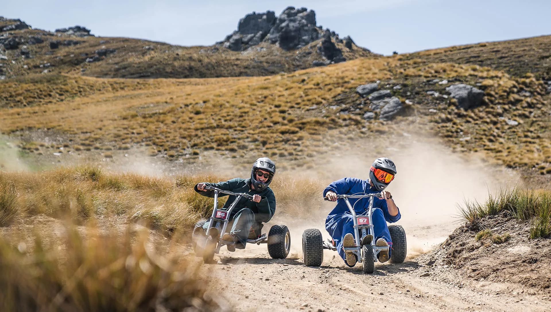 The image shows two individuals riding gravity carts down a dirt track at Cardrona Alpine Resort. Both riders are wearing helmets, with one dressed in dark green and the other in blue, racing side by side on rugged, off-road terrain. A cloud of dust follows their carts as they navigate the curvy track, set against a backdrop of rocky hills and grassy plains. The scene captures the thrill and excitement of mountain carting, showcasing the resort's adventure-filled landscape.