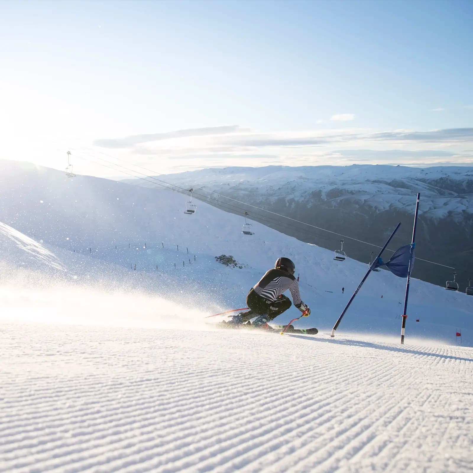 Alpine skier racing through gates on a groomed slope at sunrise, with chairlifts and mountain peaks in the background.