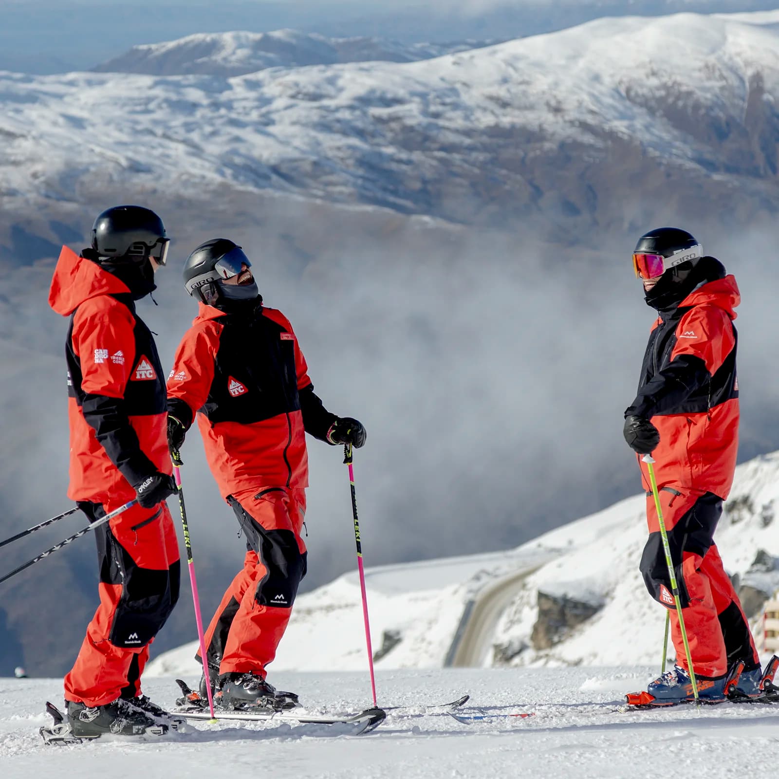 Three skiers in matching red and black outfits stand on a snow-covered mountain, holding ski poles and conversing, with misty valleys and distant snowcapped peaks in the background.