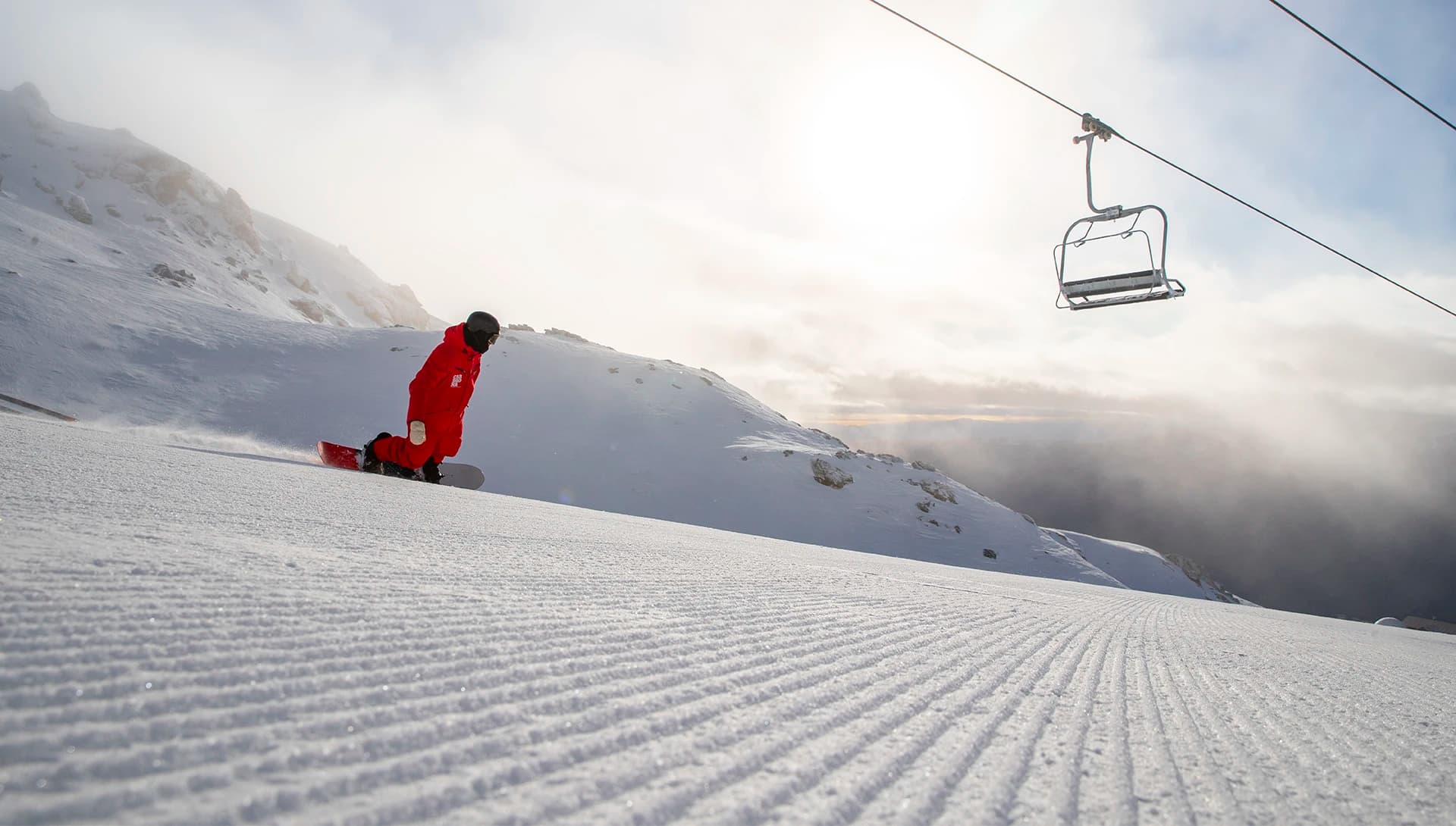 A lone snowboarder in a red jacket riding a freshly groomed snow-covered slope. The grooves in the snow lead the viewer's eye toward the distant mountains and the bright sun peeking through light clouds. A single ski lift chair hovers overhead, adding to the serene and quiet atmosphere of the scene.