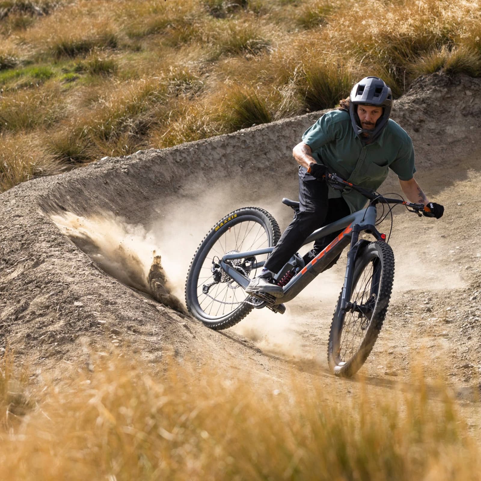 A mountain biker navigates a steep, dusty bank on a mountain bike trail. He is dressed in a teal shirt and grey helmet. As he manoeuvres the turn a cloud of dust is left behind him.