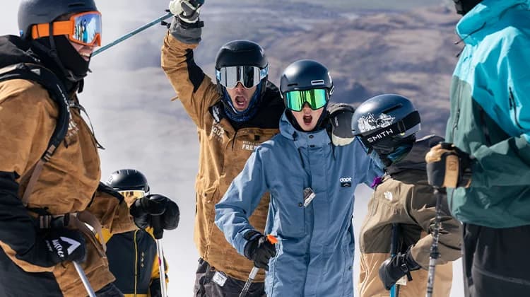 A group of skiers on a snowy mountain. In the foreground, three skiers express excitement and energy: one skier is shouting and pointing skyward, another is cheering, and a third is gesturing playfully towards the camera. The background shows a wide, expansive view of the mountain landscape under a clear sky.