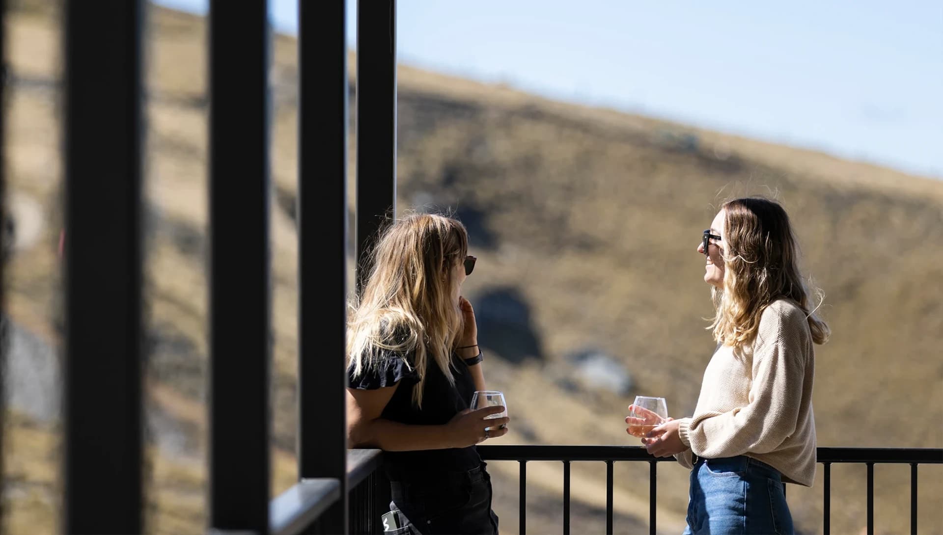 Two women enjoy a drink while standing outside on a balcony on a blue-sky summer day.