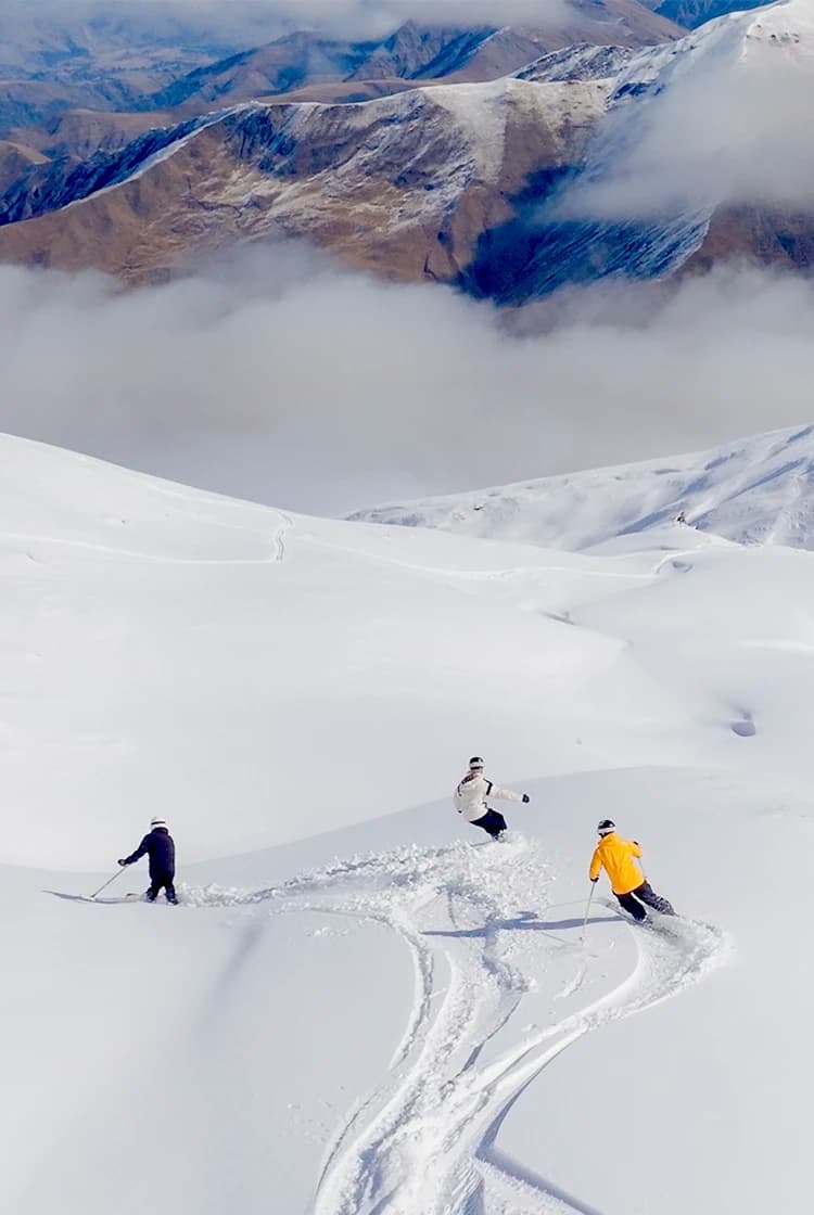 Three skiers glide down the snowy slopes of Soho Basin at Cardrona Alpine Resort, surrounded by vast, untouched powder and majestic mountain peaks. The scene is set against a backdrop of cloud-covered ridges and snow-capped peaks, offering a pristine and expansive winter landscape.