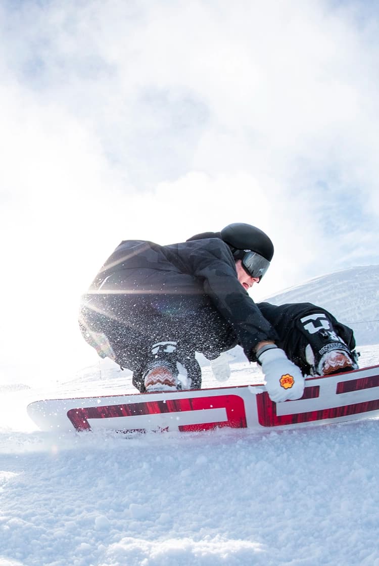 Snowboarder carving through fresh snow, with sunlight shining through mist in the background and powder kicking up from the snowboard. The rider is wearing black gear and goggles, capturing the dynamic motion and excitement of the slopes.