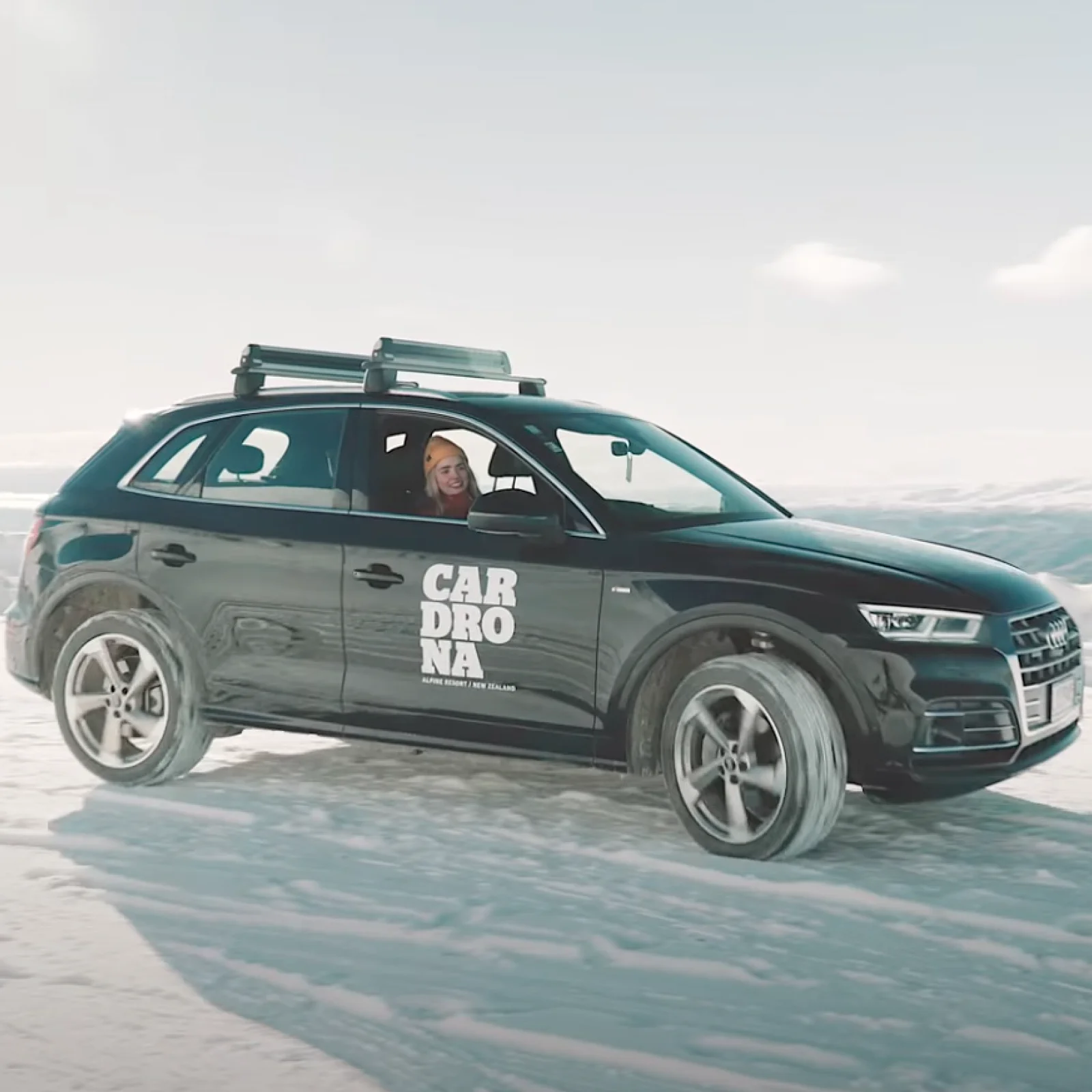 A black SUV with "Cardrona" branding drives across a snowy landscape, its tires gripping the icy terrain. A smiling driver, wearing a beanie, glances out the window.
