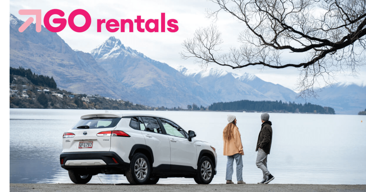 A couple stands by a white SUV parked beside a lake, with snow-capped mountains in the background and the GO Rentals logo above.