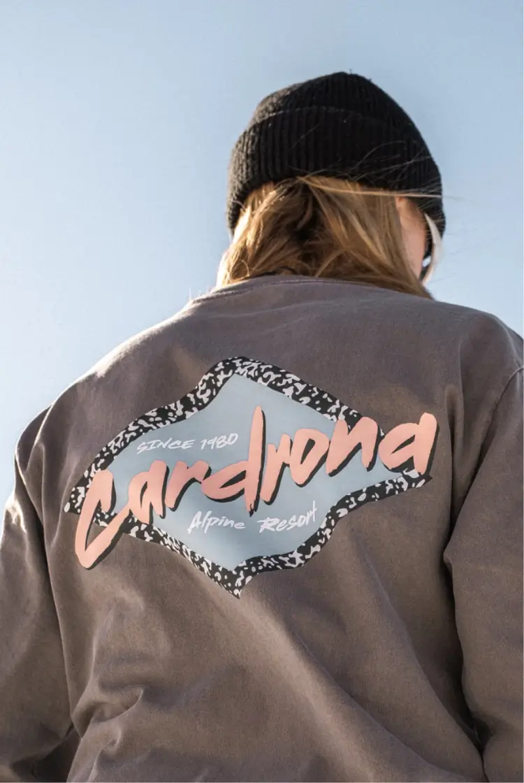 Back view of a person wearing a black beanie and a grey sweatshirt with a retro-style "Cardrona Alpine Resort" logo, standing outdoors against a clear sky.