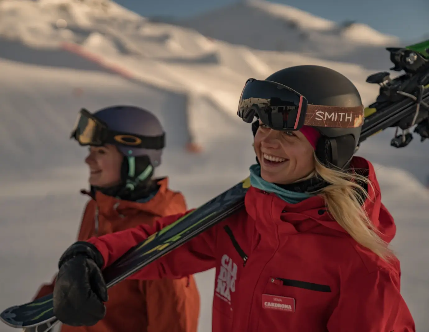 Two smiling instructors from Cardrona Snow Sports School carrying skis, dressed in branded gear, with sunny, snowy slopes in the background.