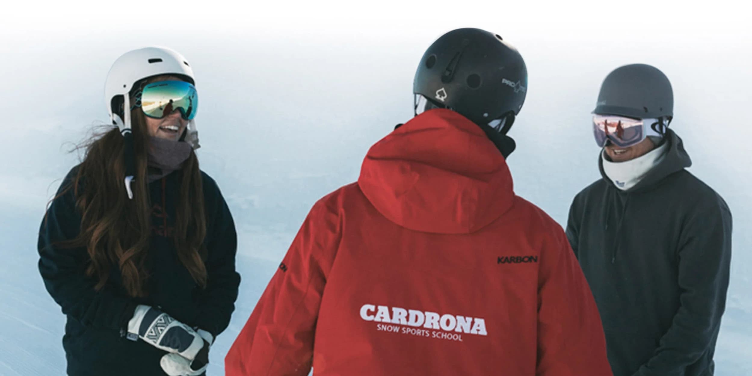 A group of three skiers or snowboarders standing together on a snowy mountain slope. All three are wearing ski gear, the person in the middle is facing away from the camera and wearing a red jacket with "CARDRONA SNOW SPORTS SCHOOL" printed on the back.