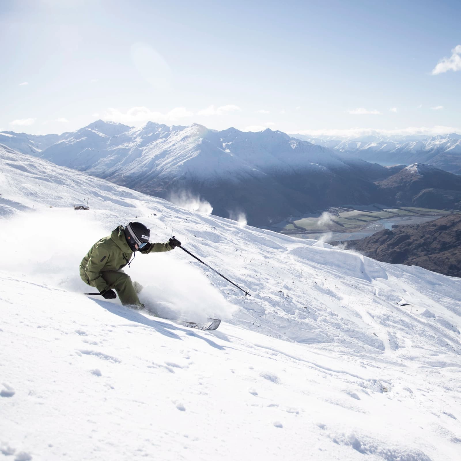 A skier makes a sharp turn through fresh powder on the slopes of Treble Cone, with expansive snow-covered mountains and a valley below under a clear blue sky.