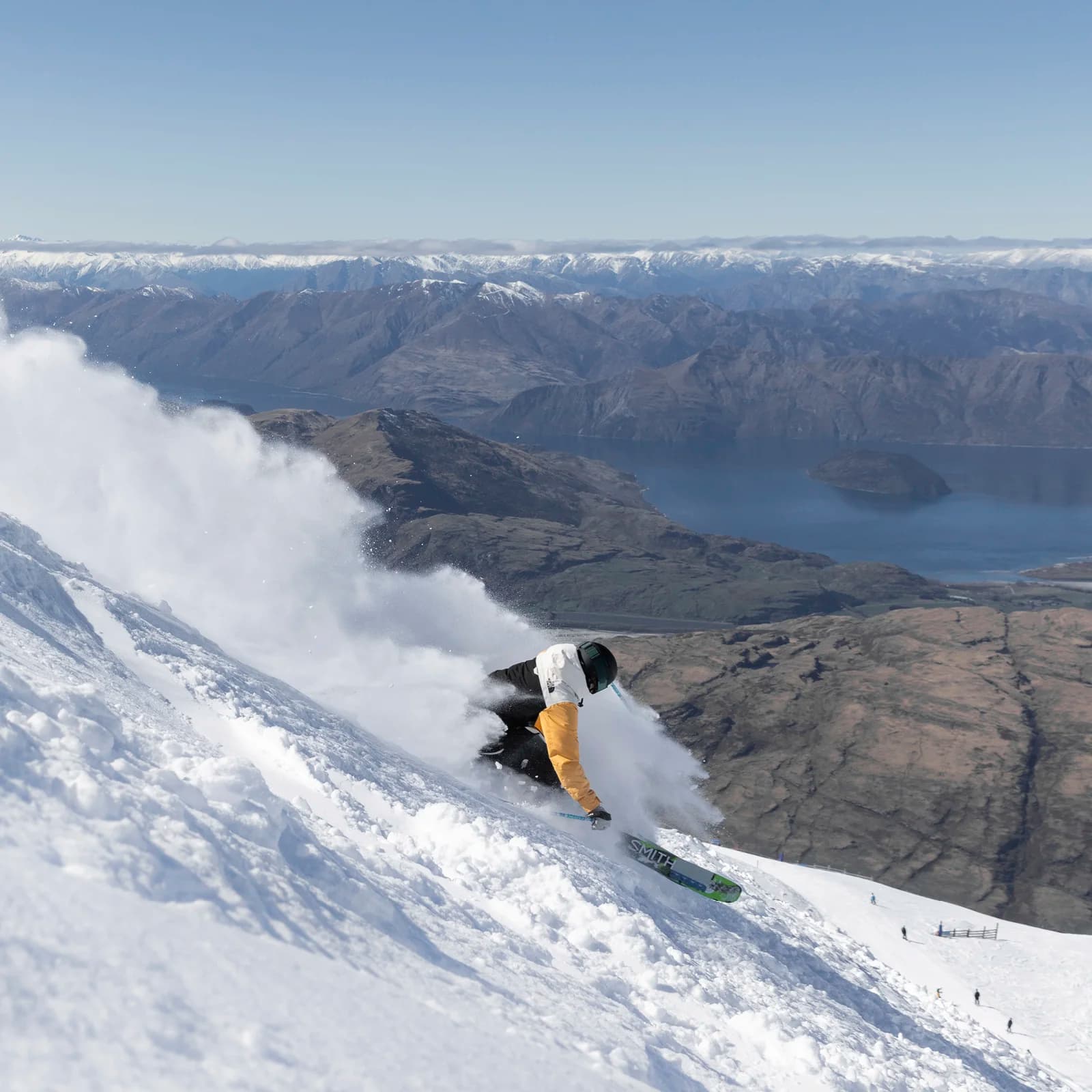 A skier carves through fresh snow on a steep slope, creating a spray of powder. The background features stunning mountain ranges and a serene lake under a clear sky.