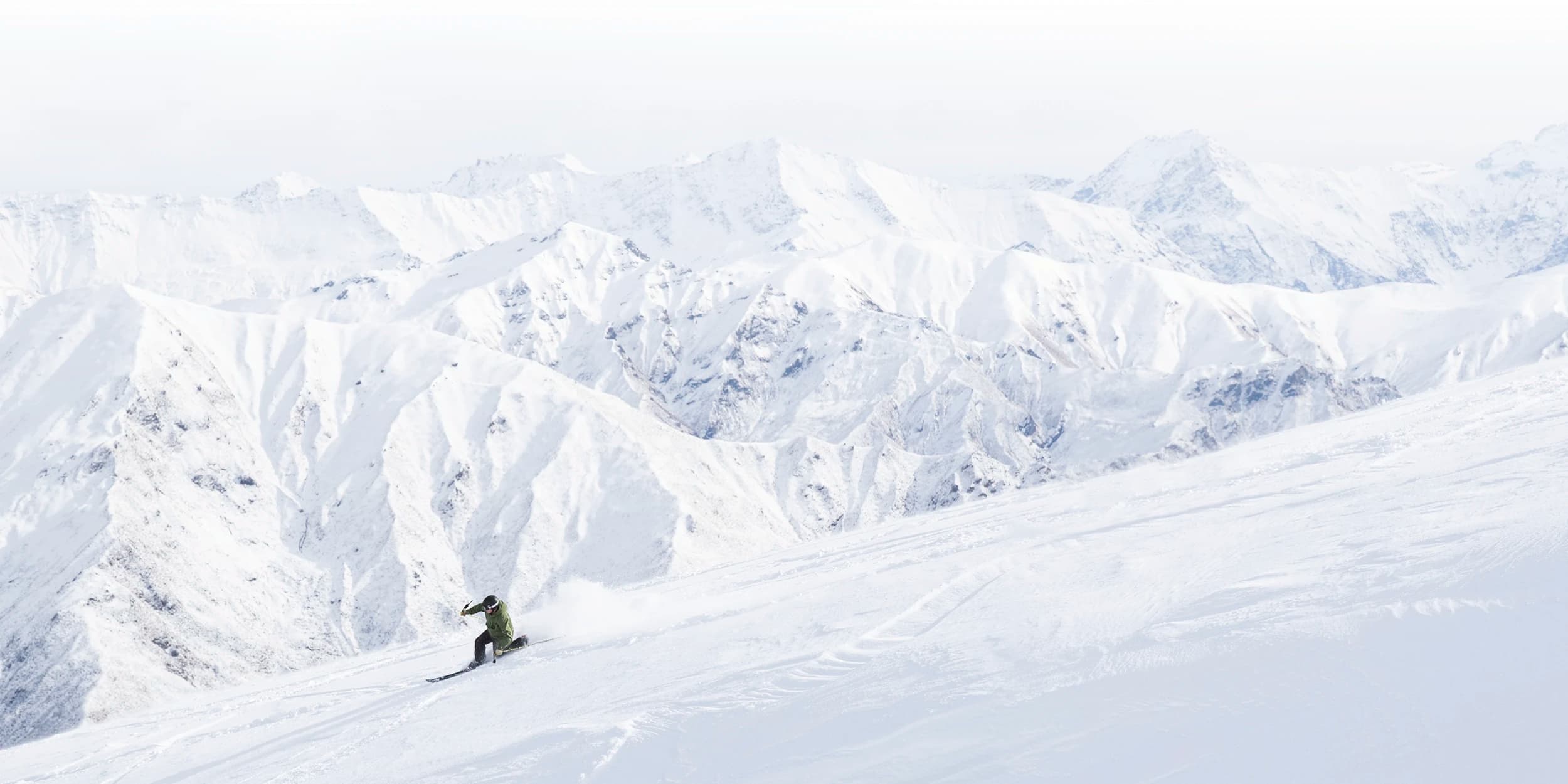 A snowboarder carves through pristine white snow on a steep slope with a dramatic backdrop of rugged, snow-covered mountain peaks under a pale sky.