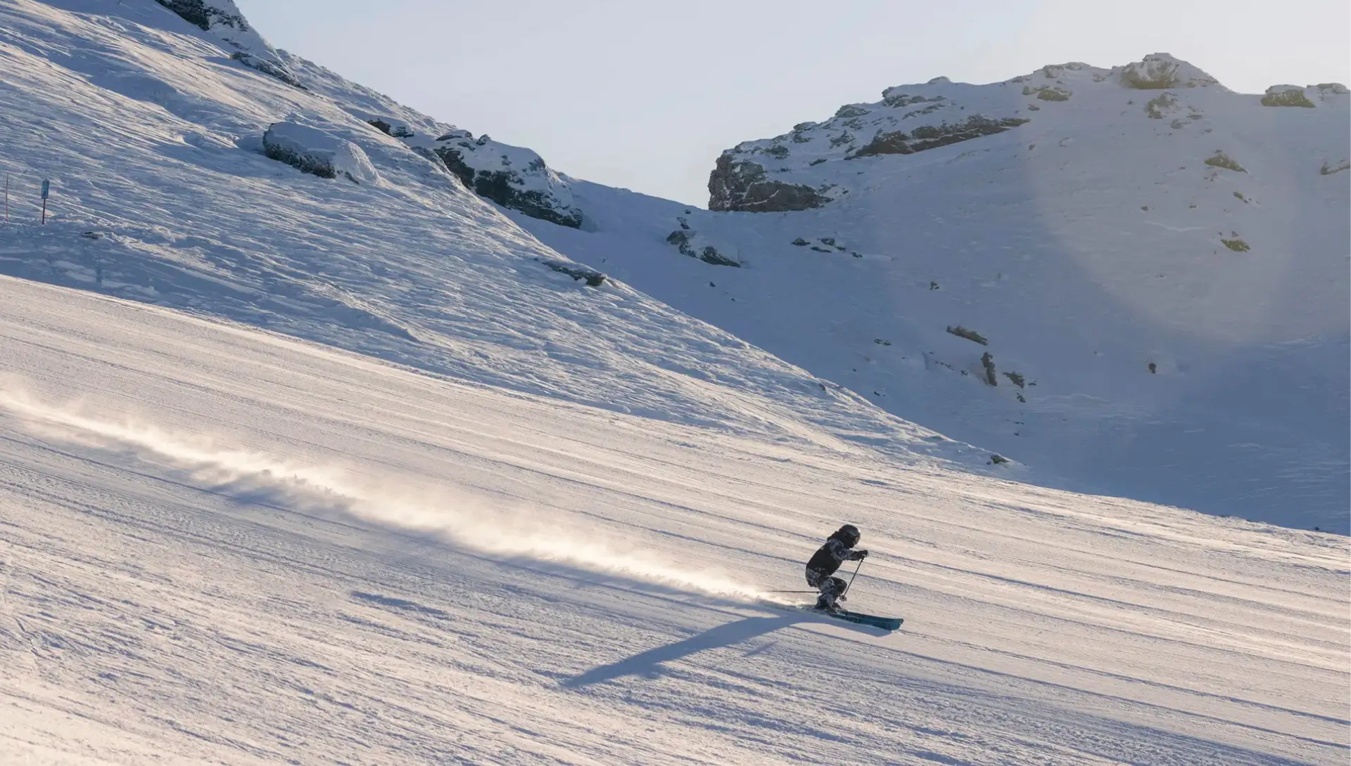 A skier glides down a wide, smooth, snow-covered slope under bright sunlight. The skier's shadow stretches out behind them as they carve gracefully, leaving a thin trail of snow dust in their wake. The surrounding mountain landscape is bathed in soft, golden light, with rocky, snow-dusted peaks rising in the background.