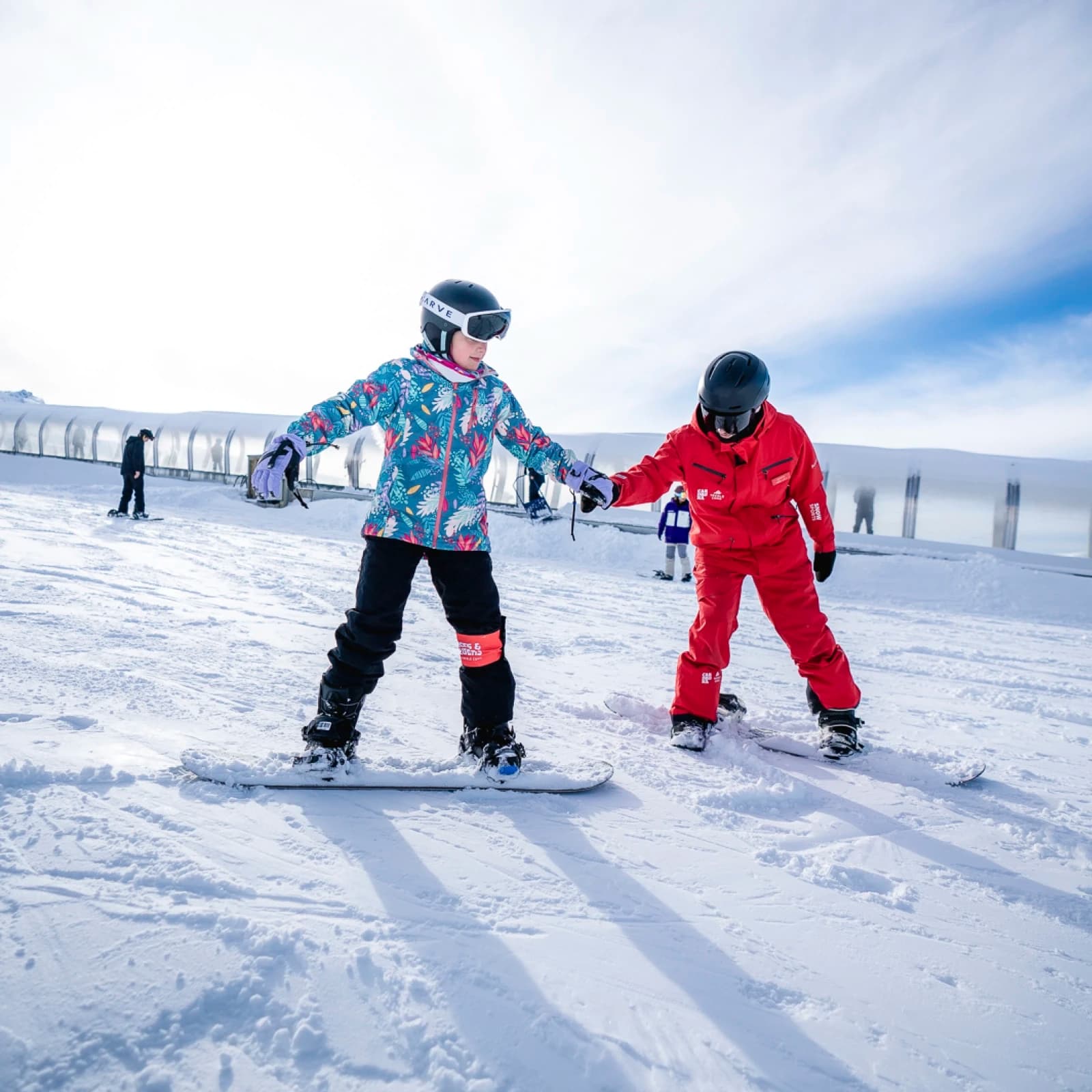 A child snowboarding with the help of an instructor on a snowy mountain. The child is dressed in a colourful, patterned winter jacket, black snow pants, gloves, and a helmet, while the instructor, wearing a bright red snowsuit and black helmet, holds the child's hand for support. Both are standing on snowboards, practicing balance on a gently sloped area. In the background, a conveyor belt-style lift and other skiers can be seen.