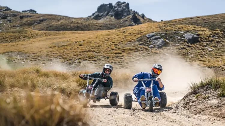 The image shows two individuals riding gravity carts down a dirt track at Cardrona Alpine Resort. Both riders are wearing helmets, with one dressed in dark green and the other in blue, racing side by side on rugged, off-road terrain.