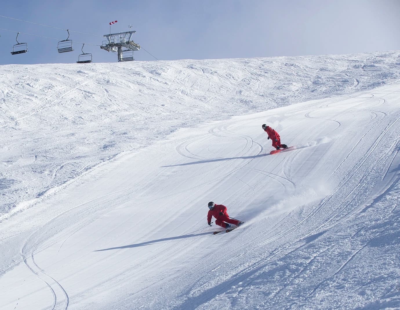 Two skiers in red gear carve smooth tracks down a pristine, snow-covered slope beneath a blue sky, with a ski lift visible in the background.