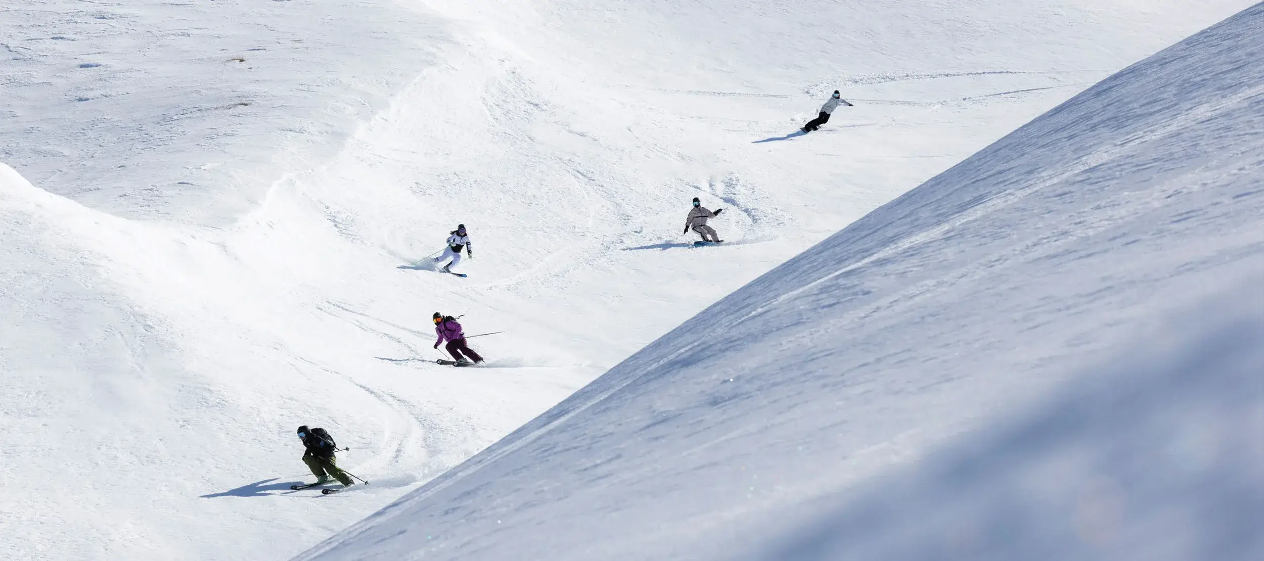 Group of skiers and snowboarders carving down a snowy slope under a clear sky.