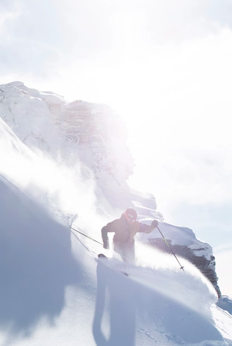 Skier carving down a steep, snow-covered mountain slope at Treble Cone Ski Field. Sunlight is illuminating the scene and casting shadows on the epic mountain landscape