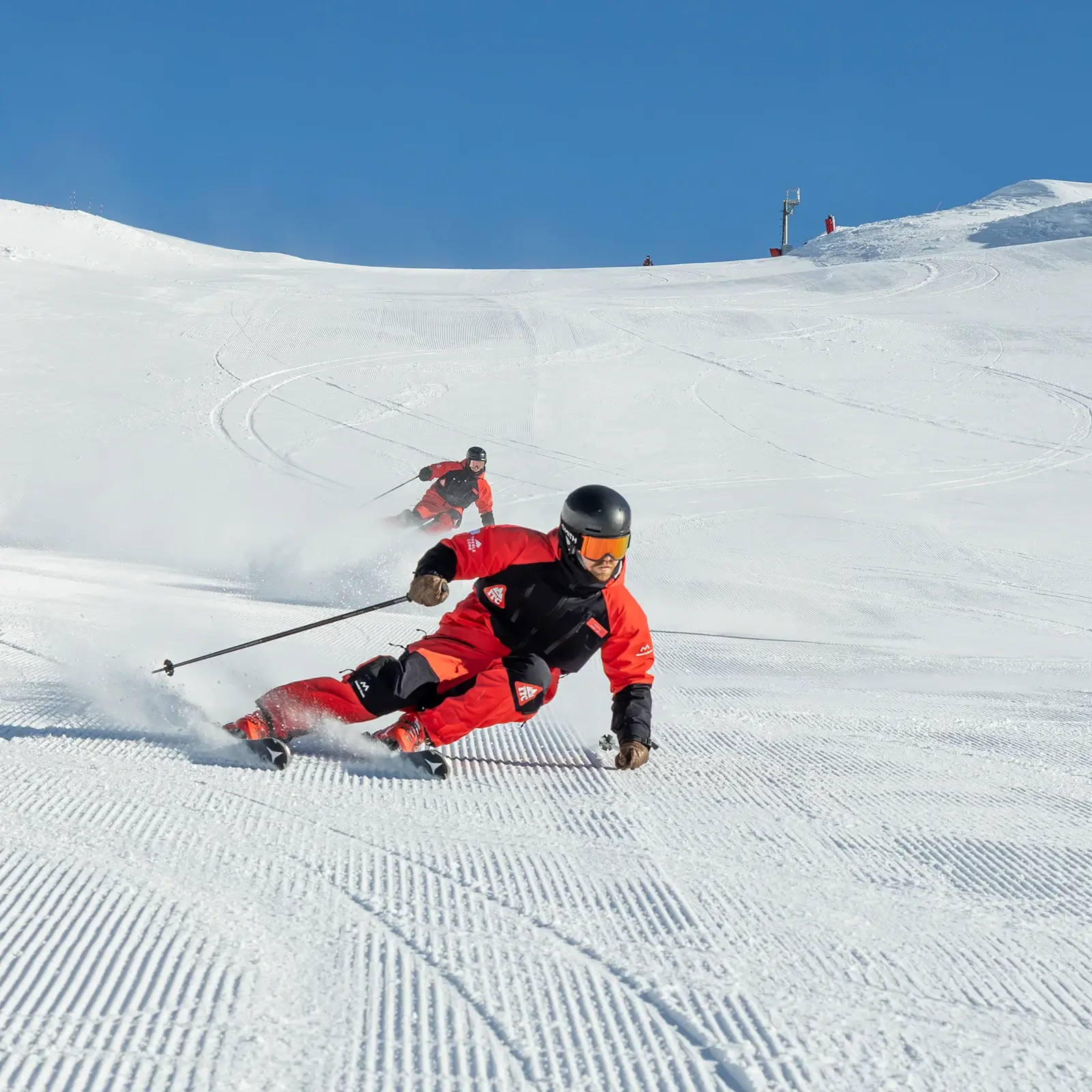 Two skiers in matching uniforms carve precise turns down a freshly groomed slope under blue skies at Cardrona Instructor Training Centre.