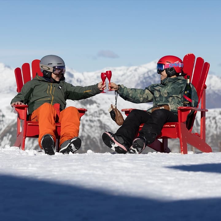 Two skiers toast on a snowy mountaintop at Cardrona in red chairs, enjoying breathtaking alpine views and a celebratory moment.
