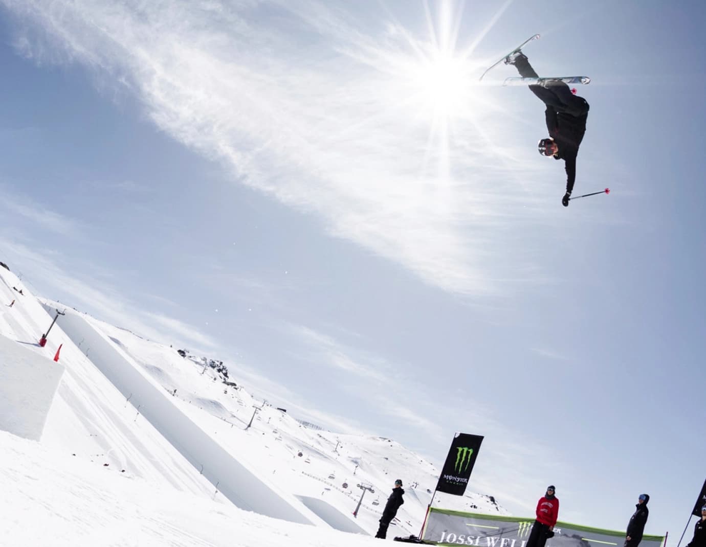 A skier performs a high-flying trick in mid-air against a bright blue sky, with spectators watching from below.