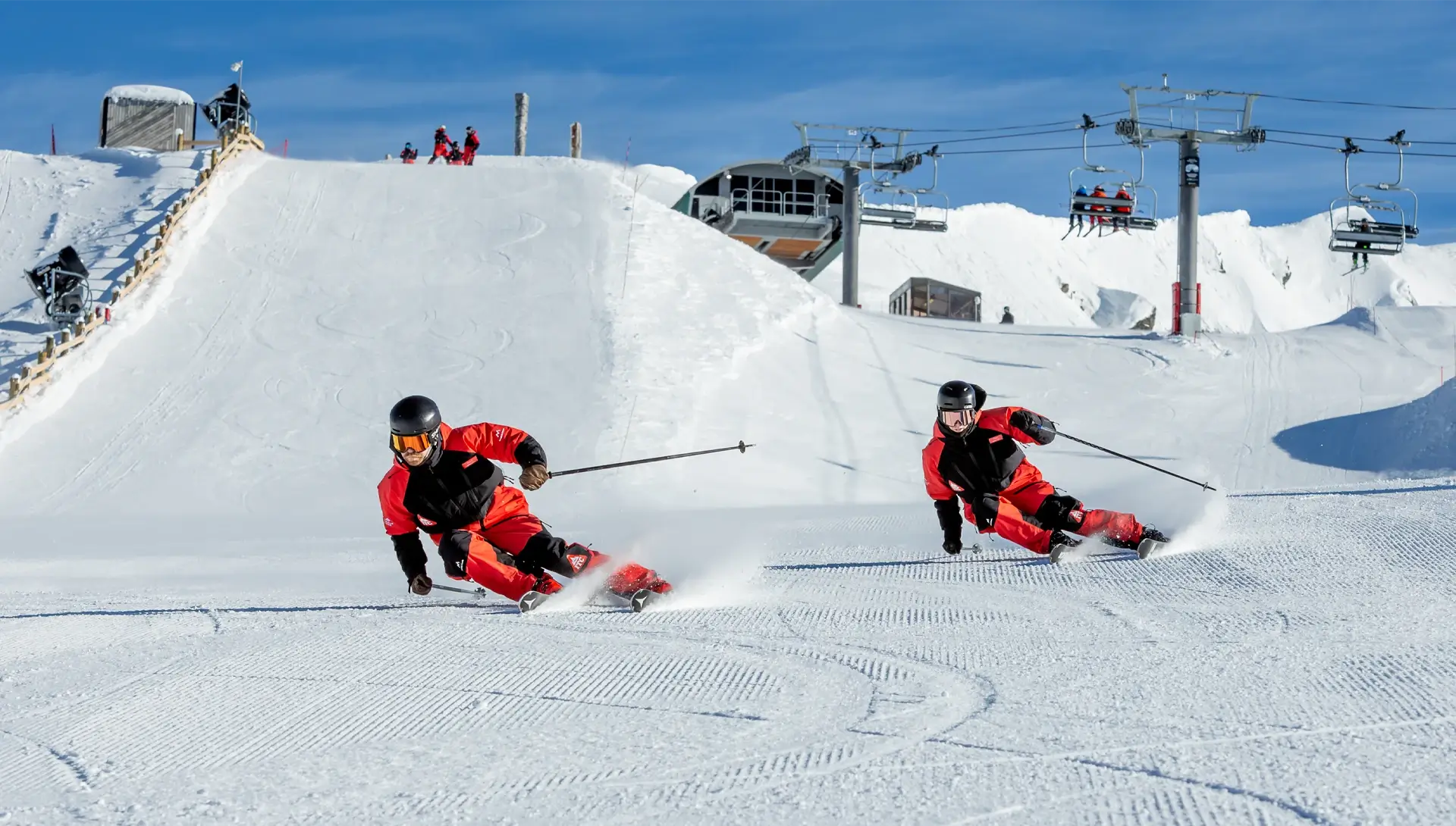 Two skiers training at Cardrona's Instructor Training Centre, carving precisely down a groomed slope beneath chairlifts on a clear, sunny day.
