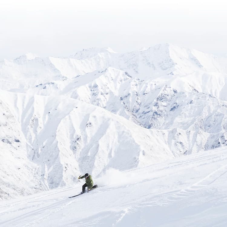 A snowboarder carves through pristine white snow on a steep slope with a dramatic backdrop of rugged, snow-covered mountain peaks under a pale sky.