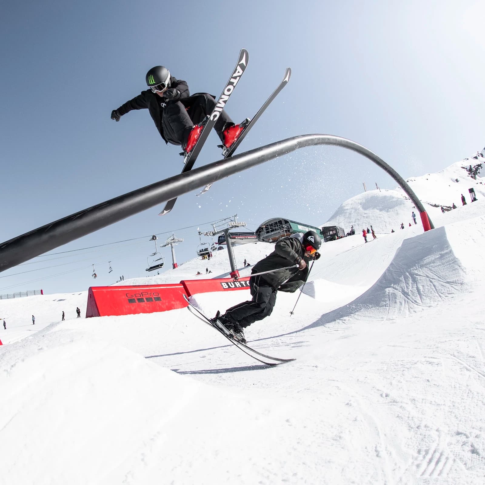 Two freestyle skiers perform tricks at the Cardrona terrain park, one jumping over a rail while the other carves below, with ski lifts and snow-covered slopes in the background on a clear day.