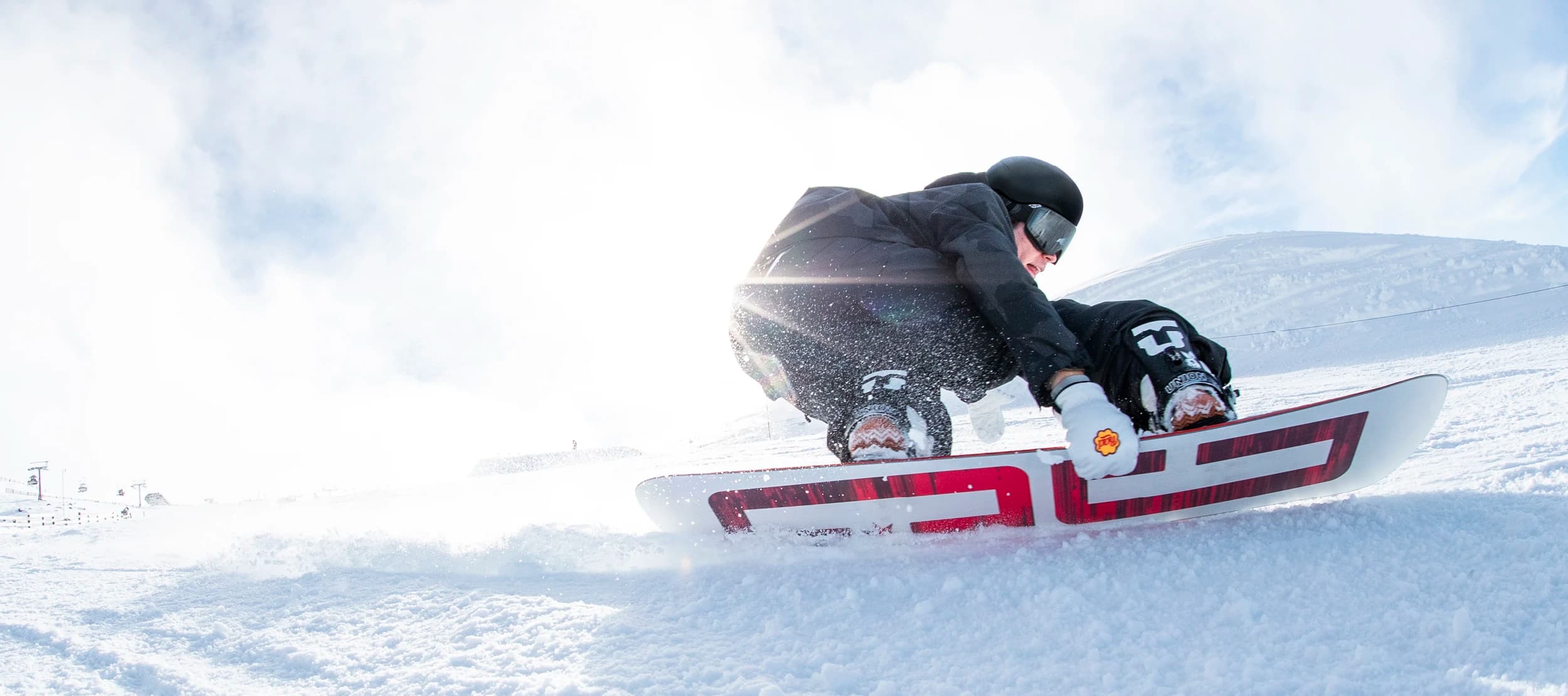 Snowboarder carving aggressively through fresh snow, with sunlight shining through mist in the background and powder kicking up from the snowboard. The rider is wearing black gear and goggles, capturing the dynamic motion and excitement of the slopes.