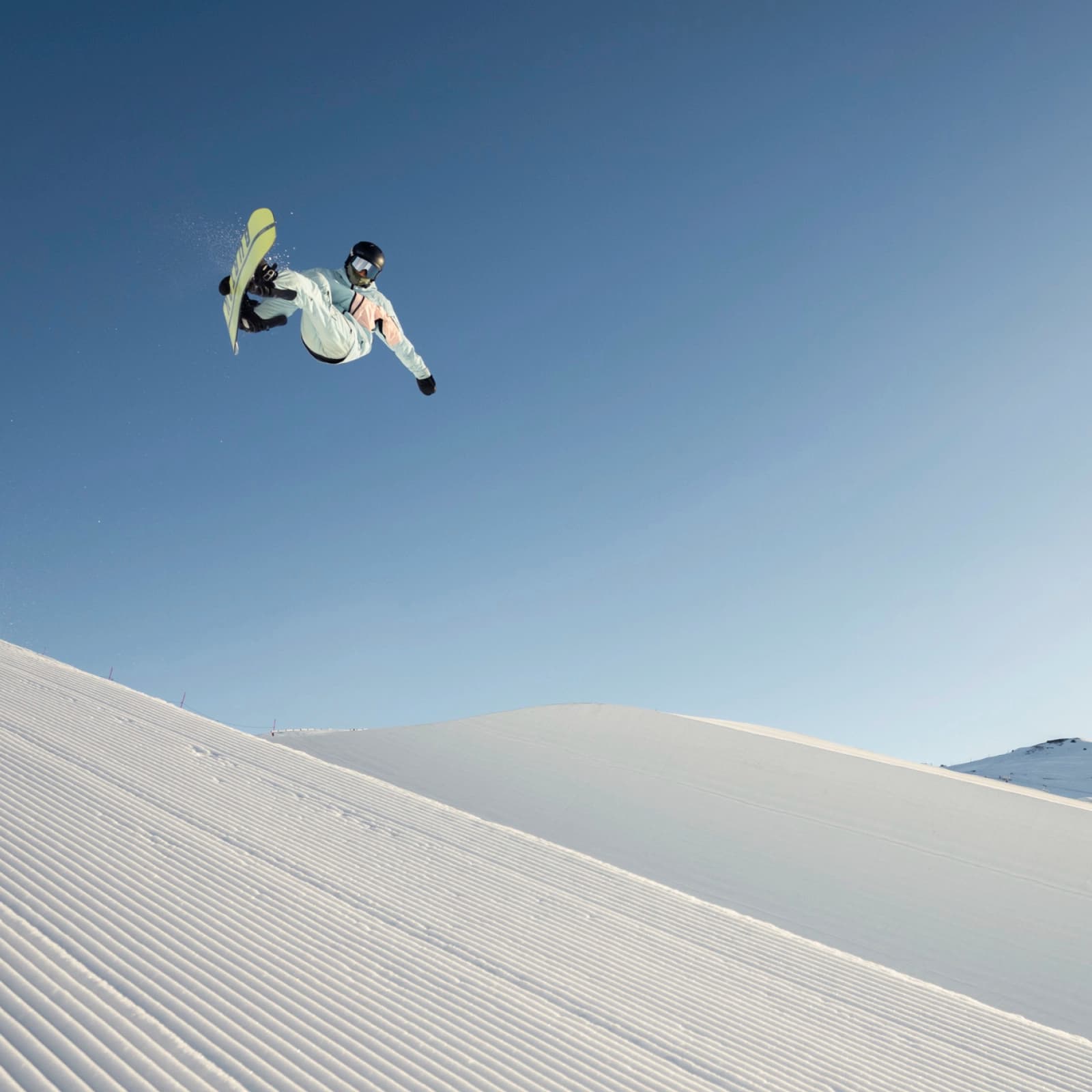 A snowboarder performing a high jump on a half-pipe, with a clear blue sky and snow-covered mountains in the background.