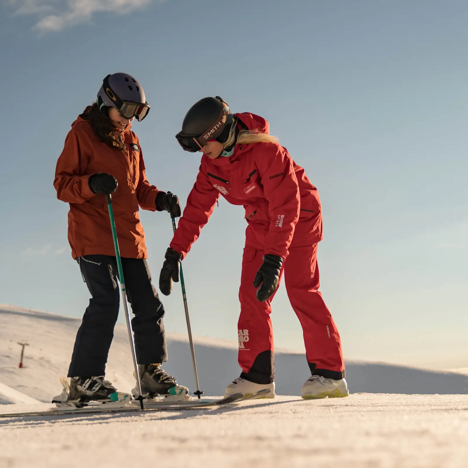 A ski instructor in red helps a beginner skier at Cardrona Alpine Resort, demonstrating technique on the snow under bright blue skies.