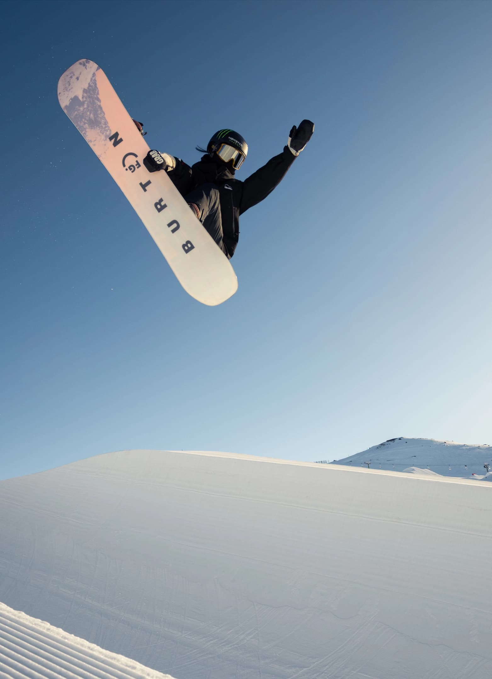 Snowboarder catching big air in the Cardrona mini pipe against a clear blue sky.