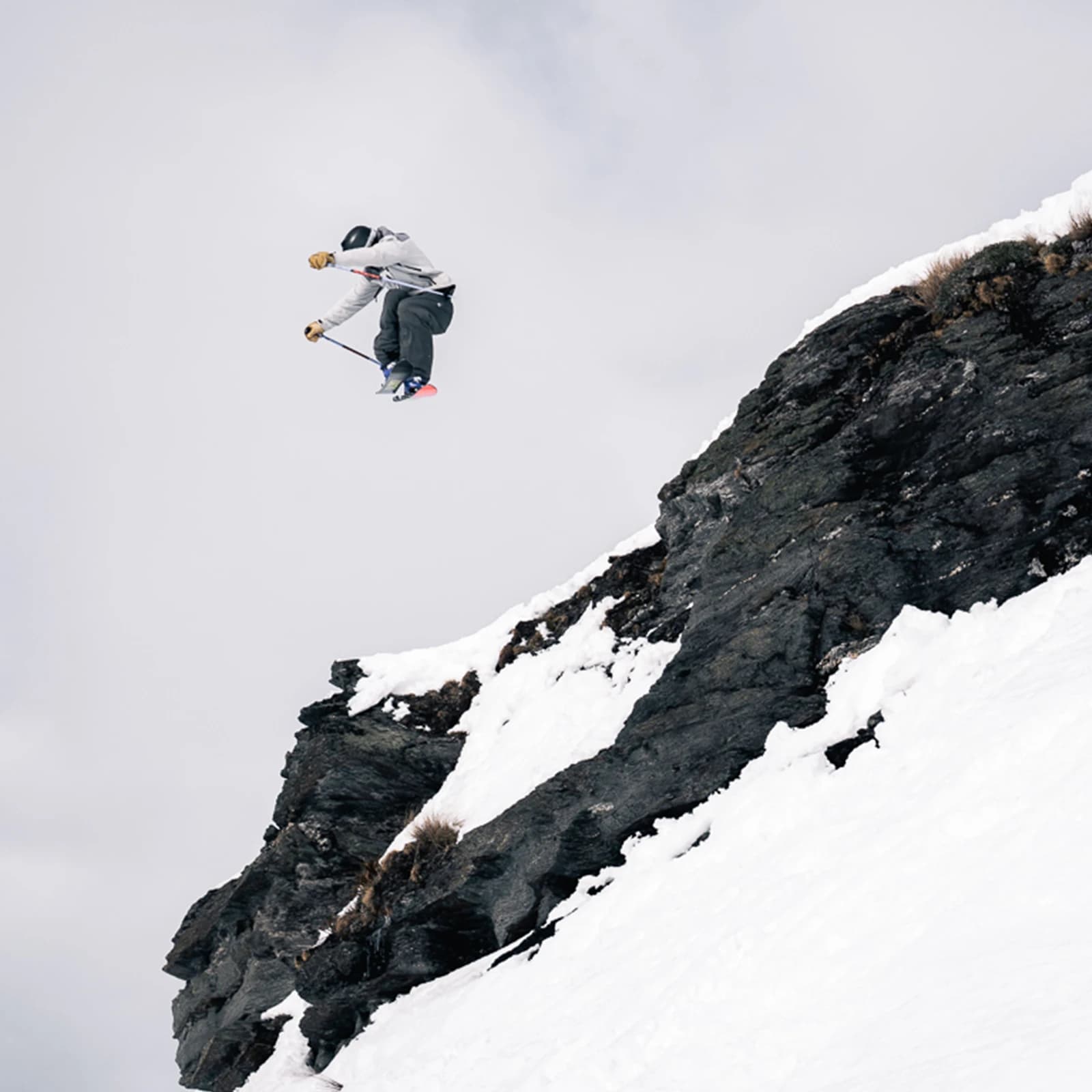 A skier mid-air, performing an impressive jump off a rocky, snow-covered cliff. The skier is wearing a grey jacket and dark pants with ski poles raised as they appear to be in full control while suspended against a cloudy sky.