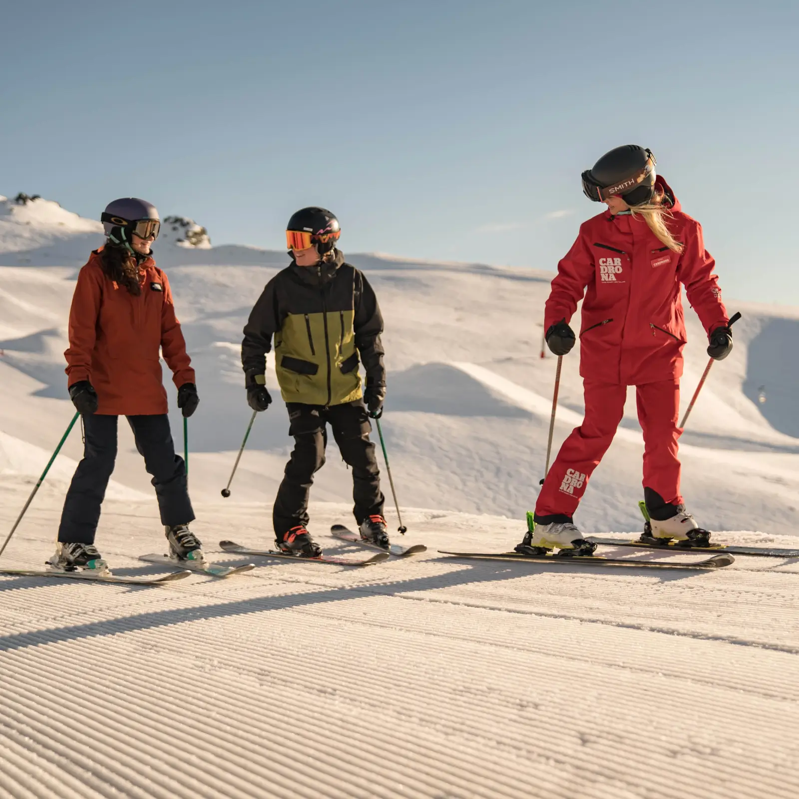 A ski instructor in red leads two skiers on freshly groomed snow at Cardrona Alpine Resort under a clear blue sky, guiding them on the slopes.