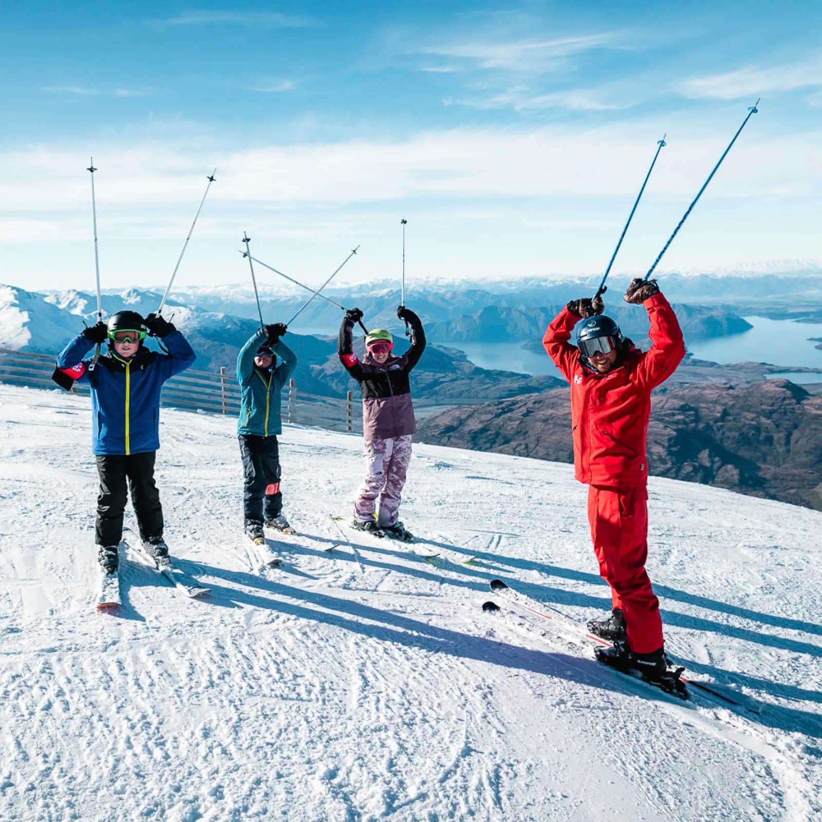 A group of skiers on a snowy mountain slope, celebrating with their ski poles raised triumphantly. From left to right, the group includes three children and one adult instructor, all wearing vibrant ski gear and helmets.