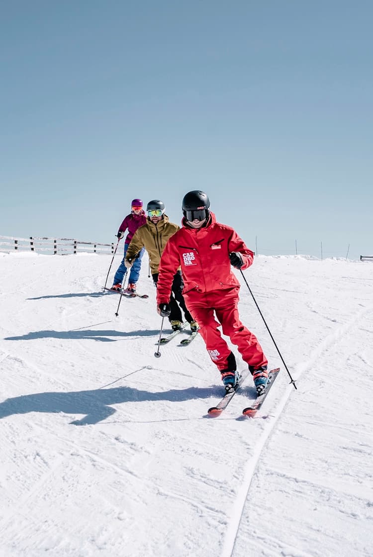 Three skiers on a snow-covered slope, following one another in a line. The skier in the front is dressed in a bright red outfit, while the two behind are in more muted colors. They are all wearing helmets and skiing under a clear blue sky, with a wooden fence lining the left side of the slope.
