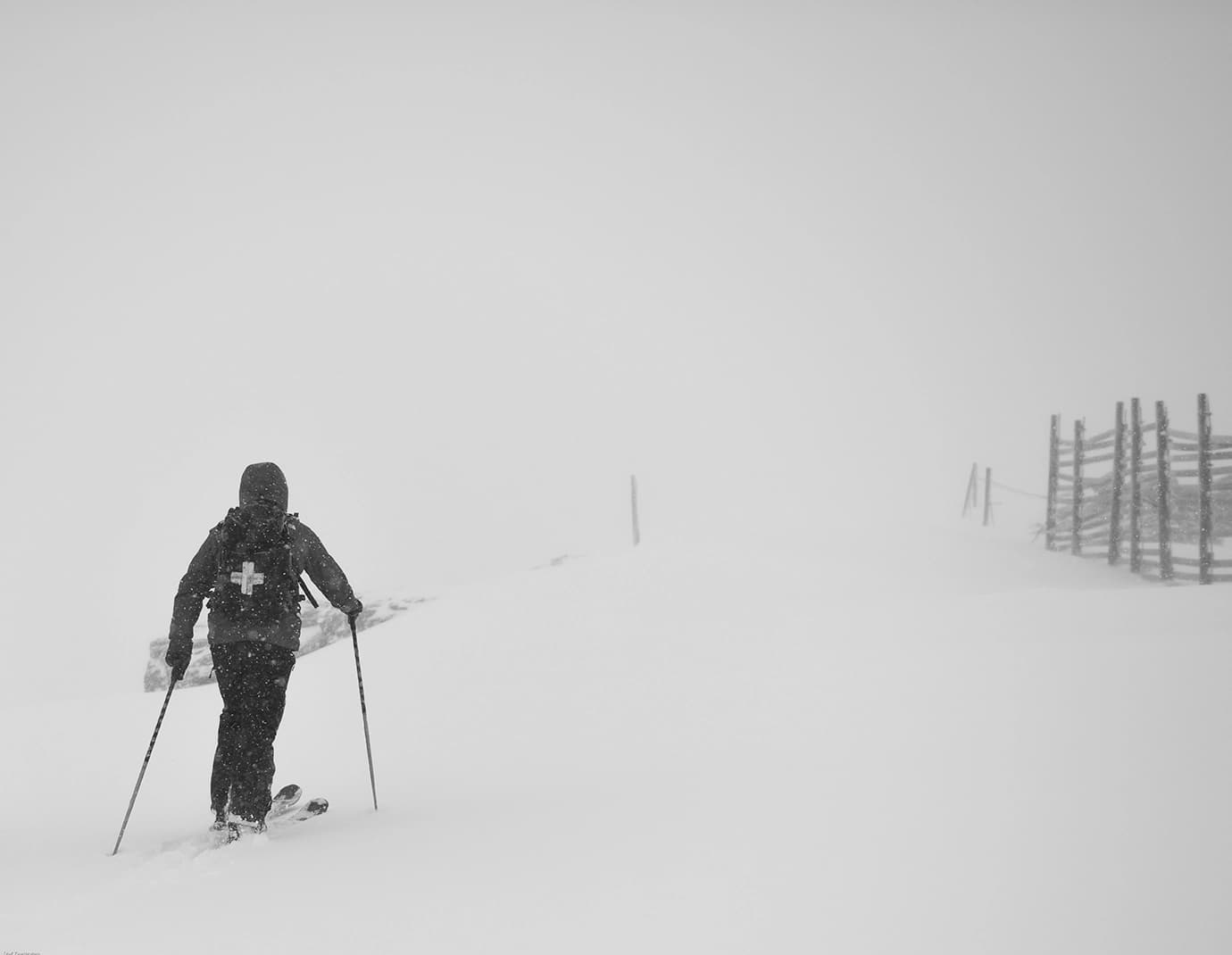A skier with a rescue backpack navigates through heavy snowfall, using poles for stability in a whiteout, near a snow-covered fence, creating a sense of isolation and challenge.