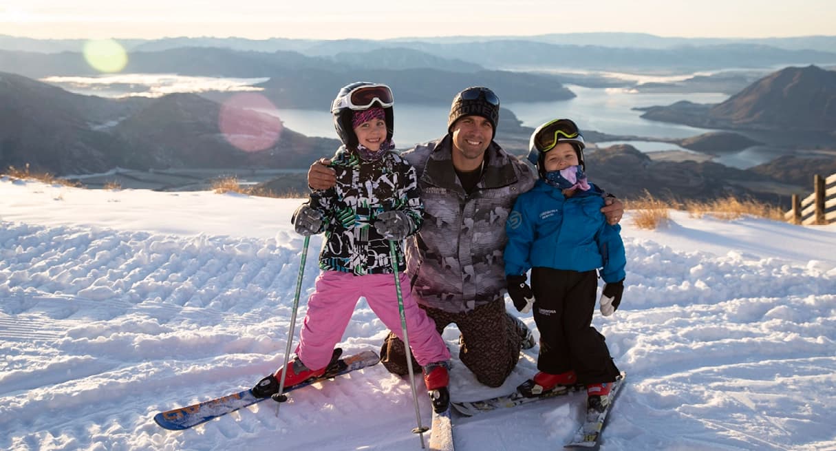 A father and two children in ski gear smile for a photo on a snowy slope at Treble Cone, with stunning views of Lake Wanaka and mountains behind them.