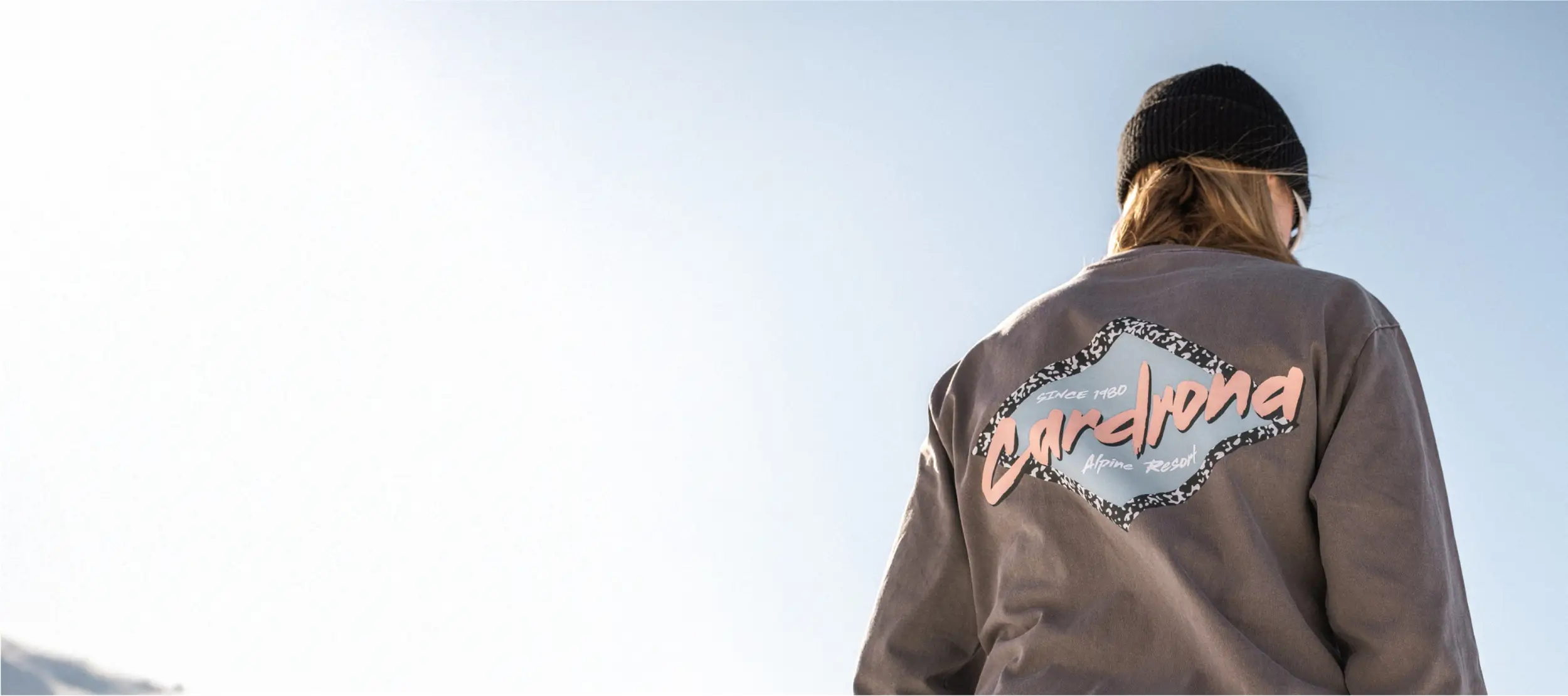 Back view of a person wearing a black beanie and a grey sweatshirt with a retro-style "Cardrona Alpine Resort" logo, standing outdoors against a clear sky.