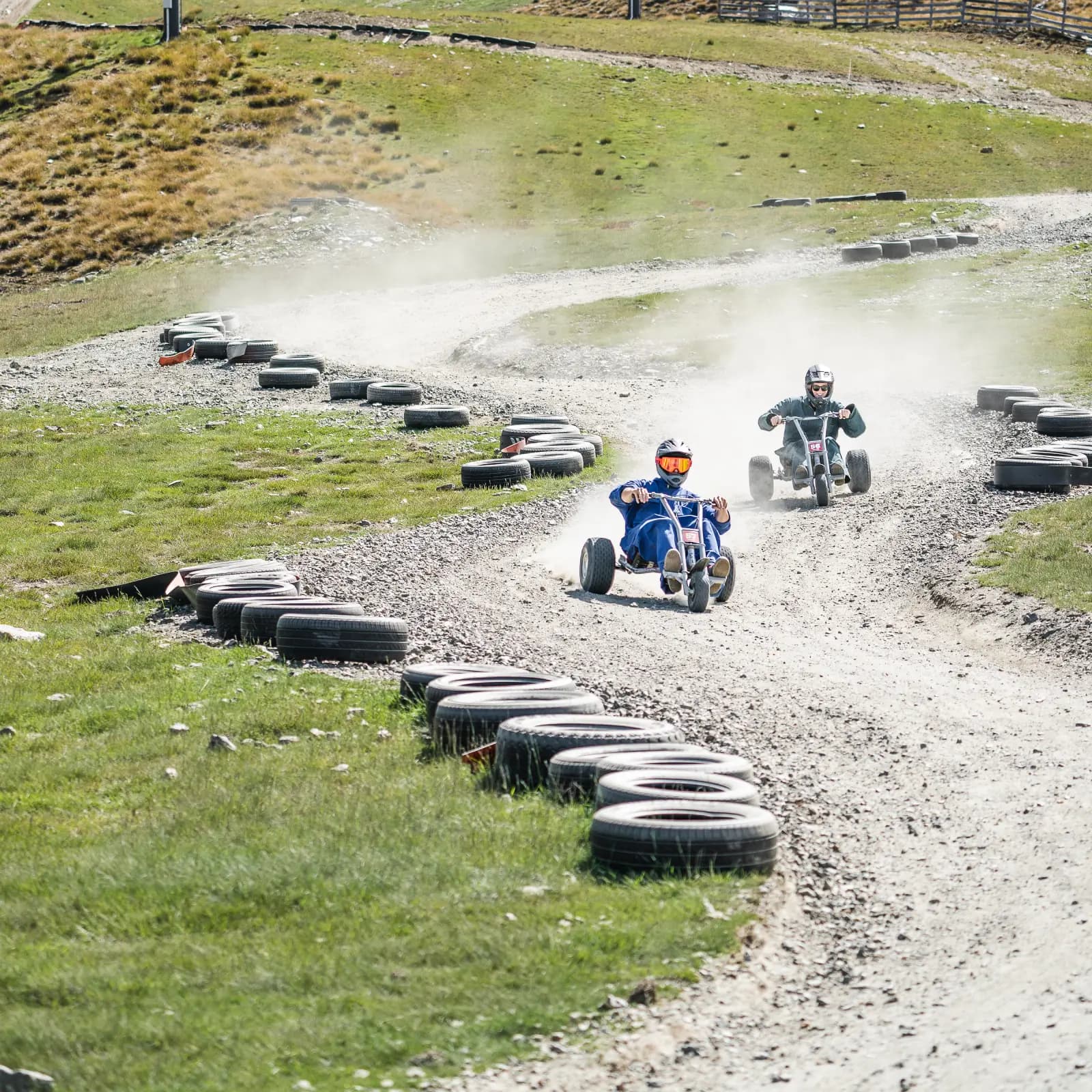 Two people racing down a winding gravel track on mountain carts, kicking up dust, with tires marking the course and grassy slopes all around.