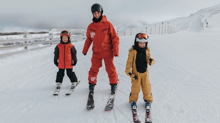 A ski instructor and two young children stand together on a snowy mountain, ready for a skiing session. The instructor, dressed in a bright red snowsuit branded "Cardrona", smiles while standing on skis. The two children, one wearing a red and black snowsuit and the other in a gold snowsuit, also have helmets and goggles. They are all positioned on a snow-covered platform with a wooden fence behind them, and the mountain's ski slopes can be seen in the distance under a cloudy sky.