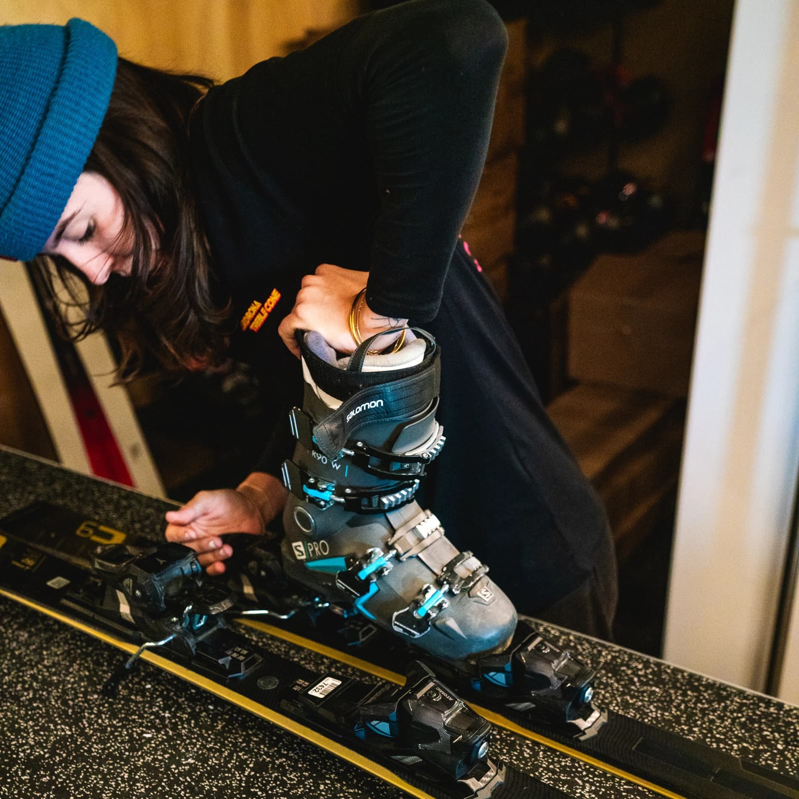 A ski technician adjusts a pair of premium ski boots on bindings, ensuring a precise fit. She works in a ski rental shop, wearing a blue beanie and dark clothing.