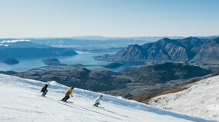 Three skiers descending a snowy slope on a clear day with a vast mountainous landscape in the background. Below the mountain range, a large blue lake is surrounded by green valleys.